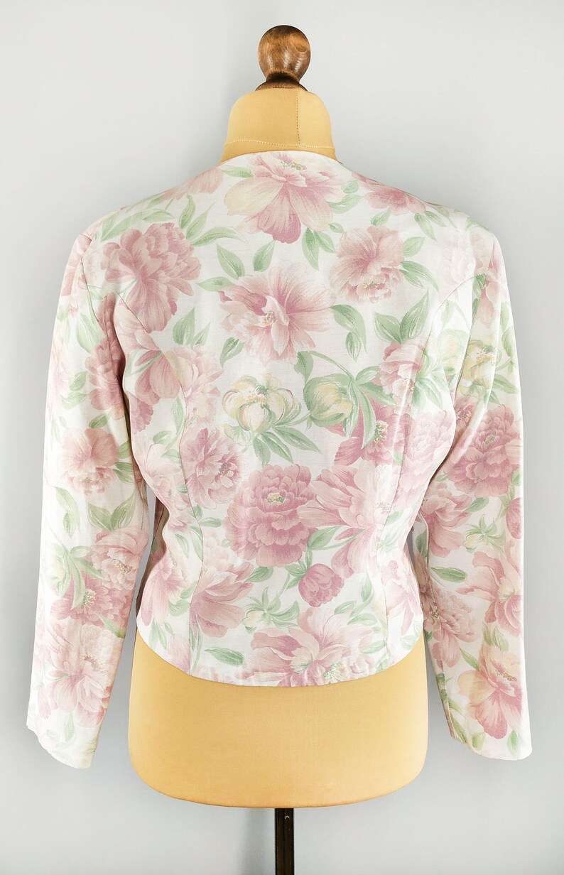 May include: A white floral print jacket with pink flowers and green leaves. The jacket has long sleeves and a fitted silhouette.