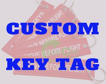 Może przedstawiać: Czerwone breloki z tkaniny z białym tekstem "Remove Before Flight". Breloki są przymocowane do srebrnych kółek do kluczy.  Custom Key Tag.