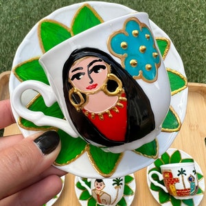 May include: A set of hand-painted ceramic coffee cups and saucers. Each cup features a different design, including a woman with a flower, a man with a palm tree, and a geometric pattern. The cups and saucers are white with green and gold accents.