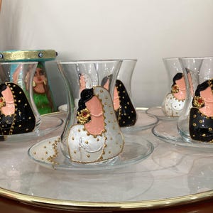 May include: Set of six clear glass teacups with painted designs of women wearing traditional clothing. The cups have gold accents and are arranged on a tray.