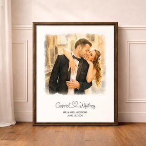 May include: A framed watercolor-style print featuring a couple embracing. The man wears a black tuxedo, and the woman is in a white dress. The print includes the names "Gabriel & Whitney" and the date "June 18, 2020". The frame is dark brown.