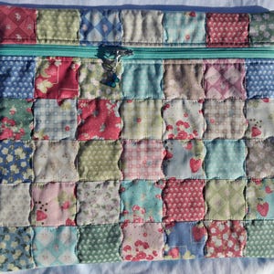 May include: A rectangular zippered pouch featuring a patchwork design of quilted fabric squares in various pastel colors, including pink, blue, green, and white. The fabric patterns include floral prints, gingham, and strawberries. A small blue butterfly charm is attached.