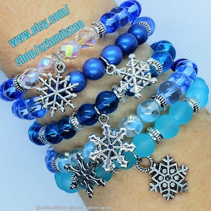 May include: Four layered bracelets with blue and aqua glass beads and silver snowflake charms. The bracelets are stacked on a person's wrist. The text "www.etsy.com/shop/azfamilyzoo" is visible in the top left corner of the image.