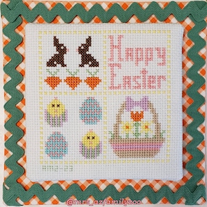 May include: A white cross-stitch embroidery with a green and orange scalloped border. The embroidery features a basket of flowers, bunnies, eggs, and the text "Happy Easter".