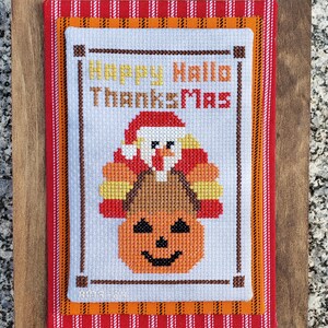 May include: Cross-stitch embroidery with a pumpkin and Santa Claus on a turkey. The text reads "Happy Hallo Thanks Mas". The embroidery is framed with a brown wooden frame and a red and orange striped mat.