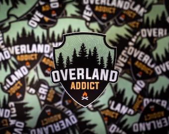May include: A green and black sticker in the shape of a shield, featuring a forest scene. The text "OVERLAND ADDICT" is in white with a yellow outline. A campfire icon is below the text.