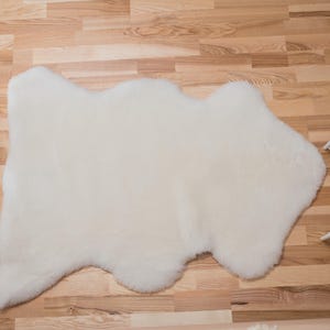 May include: A white sheepskin rug with a natural, irregular shape. The rug is laid out on a wooden floor.