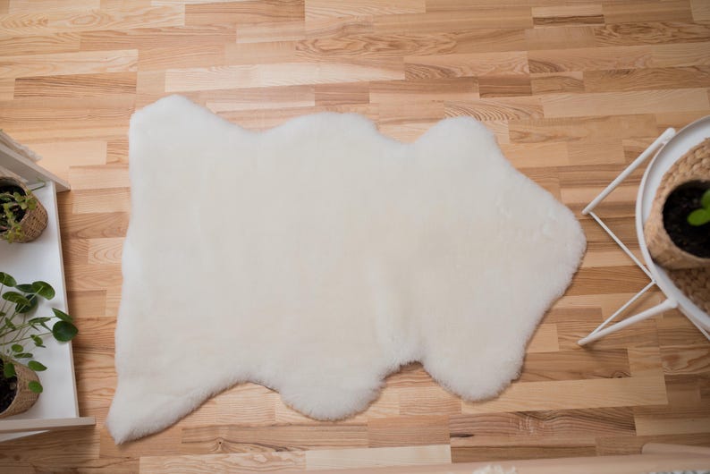 May include: A white sheepskin rug with a natural, irregular shape. The rug is laid out on a wooden floor.