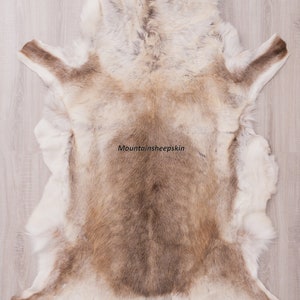 K&ouml;nnte beinhalten: Ein hellbrauner Rentierfellteppich mit wei&szlig;em Fell. Der Teppich liegt flach auf einem hellen Holzboden. Der Text "Mountainsheepskin" ist in der Mitte des Teppichs gedruckt.