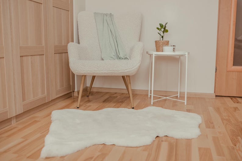 May include: A white fluffy rug with a white armchair and a small white side table with a plant in a woven basket on top. The chair has a light green throw draped over the back.