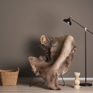 K&ouml;nnte beinhalten: Ein brauner Schaffellteppich, der &uuml;ber einen schwarzen Metallstuhl mit einer geflochtenen Holzlehne gelegt ist. Im Hintergrund steht eine schwarze Stehlampe mit goldener Akzent. Ein geflochtener Korb steht auf dem Boden.