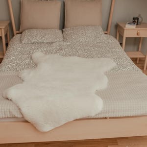 May include: A white faux fur rug on a bed with a white and gray patterned duvet cover. The bed has a wooden frame and is made up with white and gray pillows.
