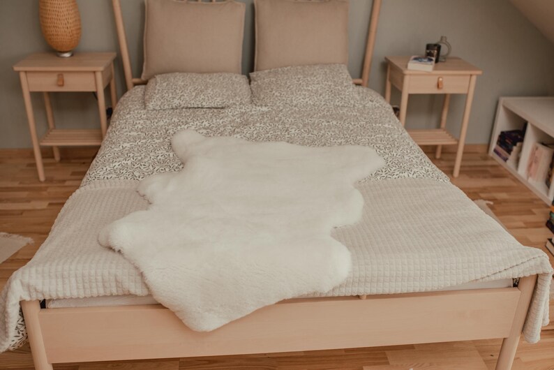 May include: A white faux fur rug on a bed with a white and gray patterned duvet cover. The bed has a wooden frame and is made up with white and gray pillows.