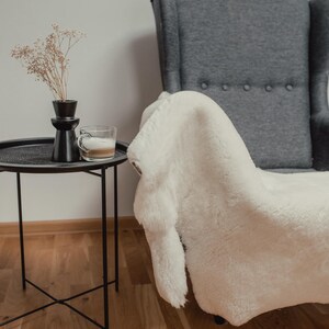 May include: A white faux fur throw draped over a gray armchair with a black side table and a vase with dried flowers.