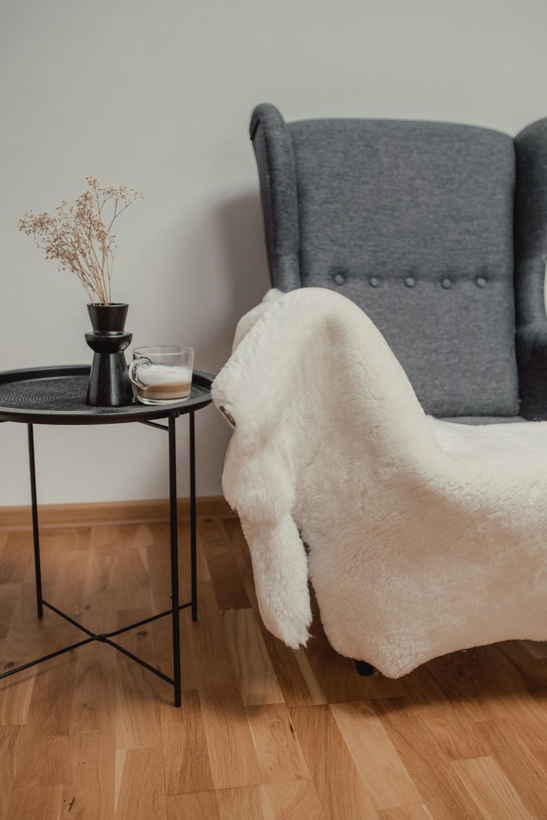May include: A white faux fur throw draped over a gray armchair with a black side table and a vase with dried flowers.