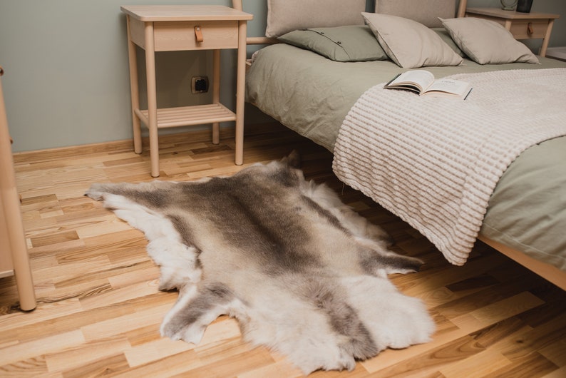 K&ouml;nnte beinhalten: Ein hellbrauner Kunstfellteppich mit wei&szlig;em Unterbauch, der auf einem hellen Holzboden in einem Schlafzimmer liegt. Ein Nachttisch aus Holz mit einer Schublade und einem Regal ist im Hintergrund zu sehen.