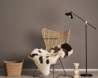 May include: A brown wicker chair with a white and brown sheepskin throw. A black floor lamp with a gold accent is in the background. The chair is on a light wood floor.