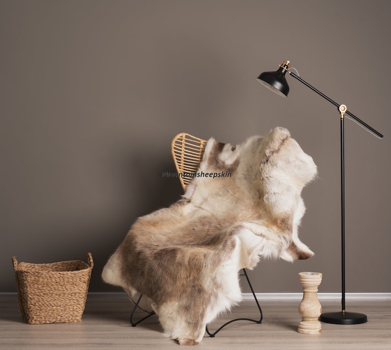 K&ouml;nnte beinhalten: Ein hellbrauner und wei&szlig;er Schaffellteppich, der &uuml;ber einen Korbstuhl mit schwarzen Metallbeinen gelegt ist. Der Teppich befindet sich in einem Wohnzimmer mit einer Stehlampe im Hintergrund.