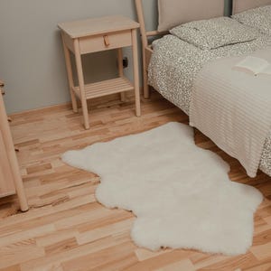 May include: A white faux fur rug on a light wood floor next to a bed with a white and gray patterned duvet cover and a white throw blanket. A small wooden nightstand with a drawer is in the background.