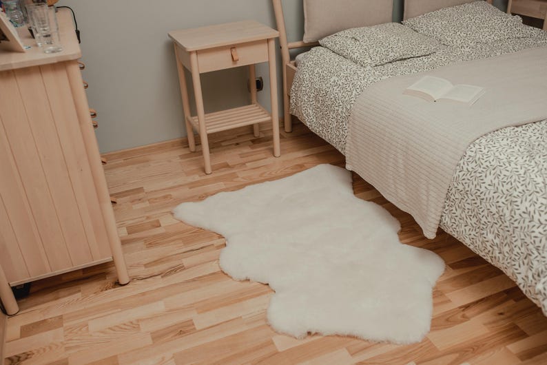 May include: A white faux fur rug on a light wood floor next to a bed with a white and gray patterned duvet cover and a white throw blanket. A small wooden nightstand with a drawer is in the background.