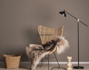 May include: A black floor lamp with a round black shade and a gold accent on the top. The lamp is next to a wicker chair with a black metal frame. A fluffy sheepskin rug is draped over the chair. A wicker basket is on the floor in the foreground.