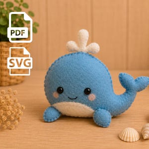 Puede incluir: Un peluche de ballena de fieltro azul con vientre blanco, un chorro de agua blanco en la parte superior y ojos de cuentas negras. La ballena tiene mejillas rosadas y una pequeña sonrisa bordada. La imagen incluye iconos de archivos PDF y SVG.