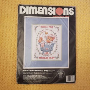 May include: A counted cross stitch pattern featuring a cartoon bear in a bathtub with bubbles. The pattern is titled "Bubble Your Troubles Away" and is from Dimensions. The pattern is 9 inches by 11 inches and includes a color accented chart.