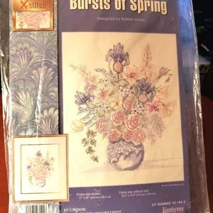 May include: A cross-stitch kit titled "Bursts of Spring" designed by Robert Jones. The kit includes 14 count white Aida fabric, pre-sorted 6 strand cotton floss, a needle, a Soft Color Graph, and instructions. The kit is for a floral design with a vase and flowers in shades of pink, purple, and white. The frame size shown is 17 inches by 20 inches, and the frame size without a mat is 12.5 inches by 15.5 inches. The kit number is 03-145 K.