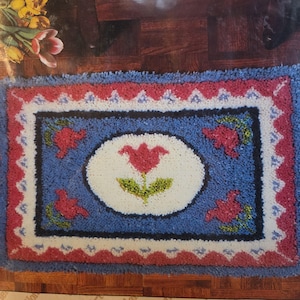 May include: A rectangular rug with a blue background and a floral design. The rug features a central oval with a red tulip and green leaves. The border is red and white with a scalloped edge.