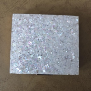 May include: Square, iridescent, decorative box with a shimmering, textured surface. The surface is covered in small, irregular pieces that reflect light, creating a sparkling effect. The box is white with a silver clasp.