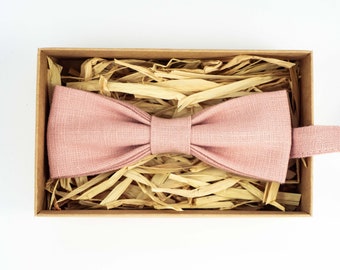 May include: A dusty rose bow tie, neatly arranged inside a small, open cardboard box filled with straw-like packing material. The bow tie is made of a textured fabric and is ready to wear.