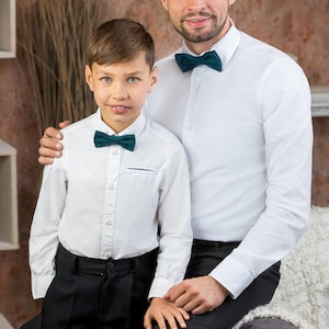 May include: A father and son wearing white dress shirts with dark green bow ties. The father has his arm around the son's shoulder. They are both smiling and looking at the camera.