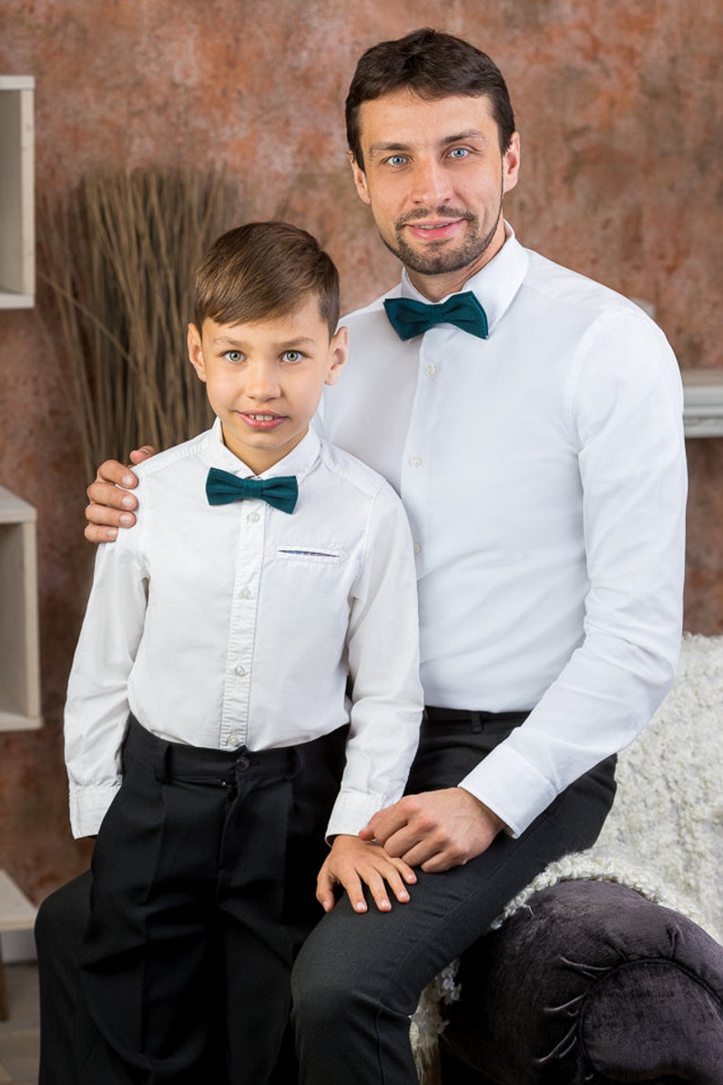 May include: A father and son wearing white dress shirts with dark green bow ties. The father has his arm around the son's shoulder. They are both smiling and looking at the camera.