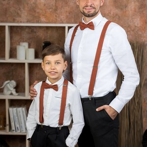May include: Two people wearing white shirts with brown suspenders and bow ties. The person on the left is a child and the person on the right is an adult. They are both smiling and looking at the camera.
