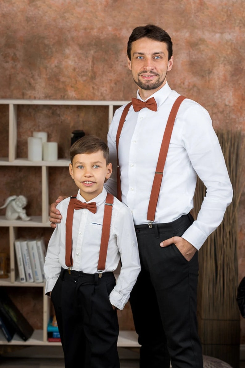May include: Two people wearing white shirts with brown suspenders and bow ties. The person on the left is a child and the person on the right is an adult. They are both smiling and looking at the camera.