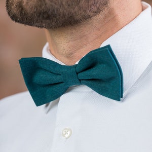 May include: A dark teal bow tie made of a textured fabric. The bow tie is tied and worn by a person wearing a white collared shirt.