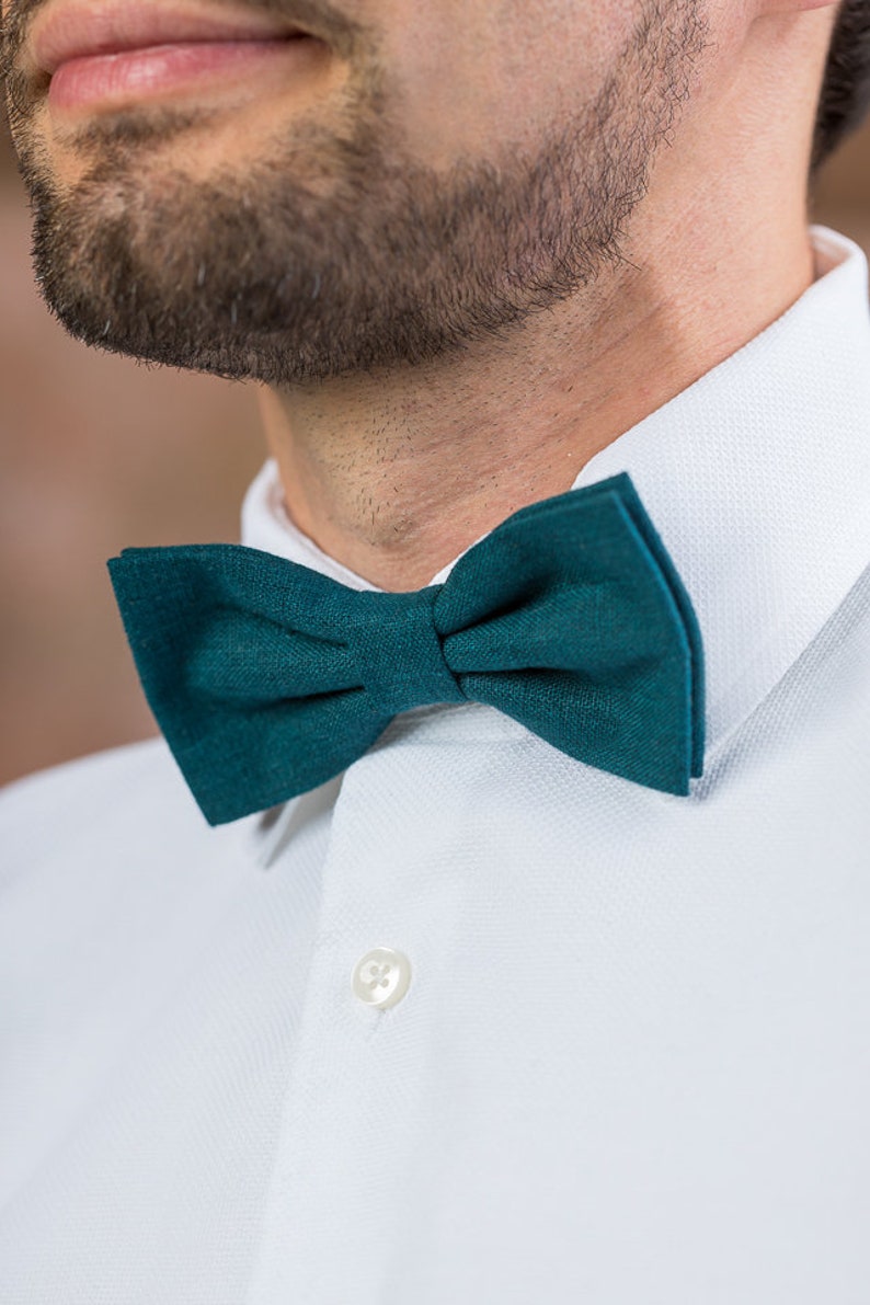May include: A dark teal bow tie made of a textured fabric. The bow tie is tied and worn by a person wearing a white collared shirt.