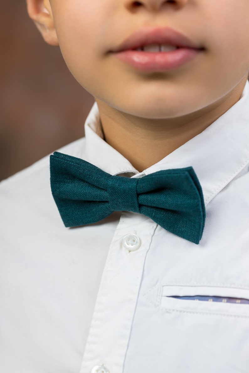 May include: A close-up of a boy wearing a white button-down shirt with a dark green bow tie.