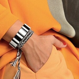 Puede incluir: Una persona lleva una pulsera de rayas blancas y negras con una pulsera tejida en blanco y negro en la muñeca. La persona lleva una sudadera naranja brillante y pantalones naranja.