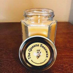 May include: A small, clear glass jar candle filled with yellow wax. The lid is off, revealing the candle inside. The lid is black with a circular label that reads "The Queens Beeswax" and features a cartoon bee.