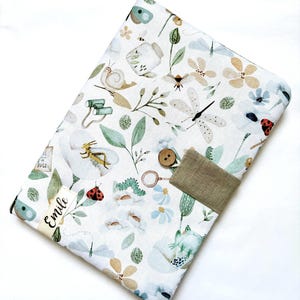Peut inclure: Une housse en tissu blanc avec un motif fantaisiste d'insectes et de fleurs. Le motif comprend des illustrations d'insectes, de fleurs et de feuilles aux couleurs douces. Un bouton marron et une sangle en tissu beige fixent la housse. Le nom "Emile" est imprimé sur une petite étiquette.