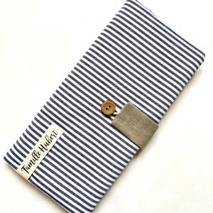 May include: A rectangular case with navy and white stripes, secured with a beige fabric loop and a wooden button. A small label with the text "Famille Hubert" is attached to the case. The case is likely for storing items.