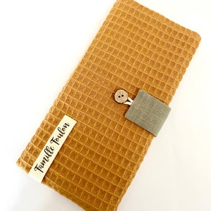 May include: A brown waffle weave fabric wallet with a gray linen strap and a wooden button. The wallet has a white label with the text "Famille Toulon" printed on it.