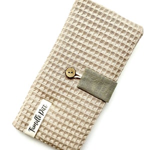 May include: A beige waffle weave fabric pouch with a green linen flap and a wooden button closure. The pouch has a white label with the text "Famille Pitt" in black cursive.