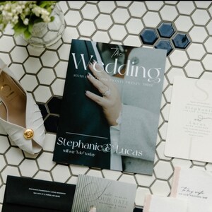 Könnte beinhalten: Ein Hochzeits-Programm mit einem dunkelblauen Umschlag, der ein Foto von den Händen eines Paares mit einem Ring am Finger der Frau zeigt. Der Umschlag des Programms trägt den Text "The Wedding" und "Stephanie & Lucas" in weißer Schrift. Das Programm liegt auf einer weißen gekachelten Oberfläche mit weißen Hochzeitsschuhen und anderen Hochzeitspapieren.