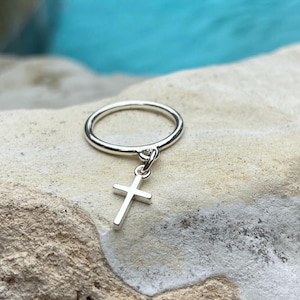 Puede incluir: Un anillo de plata con un pequeño dije de cruz de plata colgando de él. El anillo está sobre una roca marrón claro.