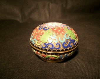 May include: A small, round, decorative box with a lid. The box is made of metal and is decorated with a colorful floral pattern. The box is about 2 inches in diameter.