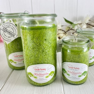 Op de afbeelding: Drie glazen potten met groene kaarsen erin. De kaarsen zijn gelabeld met 'Candle Factory' en 'Waldmeister' met de tekst 'frische Waldmeisterblüten' eronder. De potten hebben een doorzichtig glazen deksel.