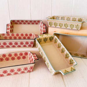 Könnte beinhalten: Eine Sammlung rechteckiger Keramik-Backformen. Einige sind mit roten Blumenmustern verziert, andere mit grünen Blattmotiven. Jede Form hat Griffe und einen cremefarbenen Innenraum.