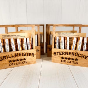 Op de afbeelding: Twee houten kisten met de woorden "Grillmeister De Luxe" en "Sterneküche De Luxe" erop gedrukt. Elke kist bevat kleine glazen potjes met verschillende dipsmaken. De kisten zijn ideaal voor het opbergen en presenteren van kleine voorwerpen.